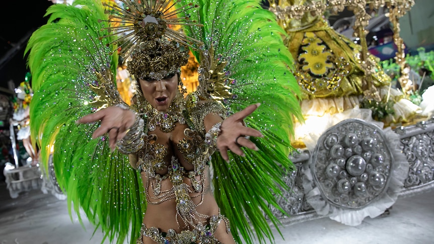 a dancer in a gold outfit adorned with green feathers holds out their hands toward the camera as they dance in front of a float