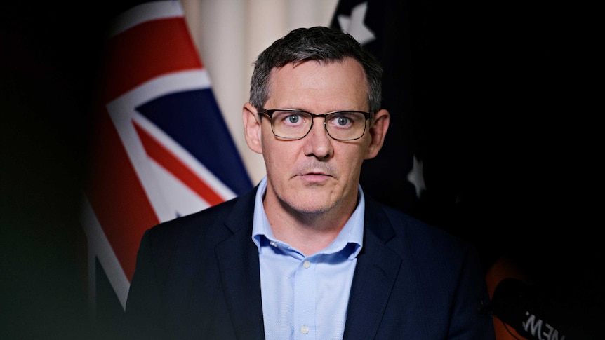 Chief Minister Michael Gunner is standing in front of a microphone with a serious expression. Behind him is the Australian flag.