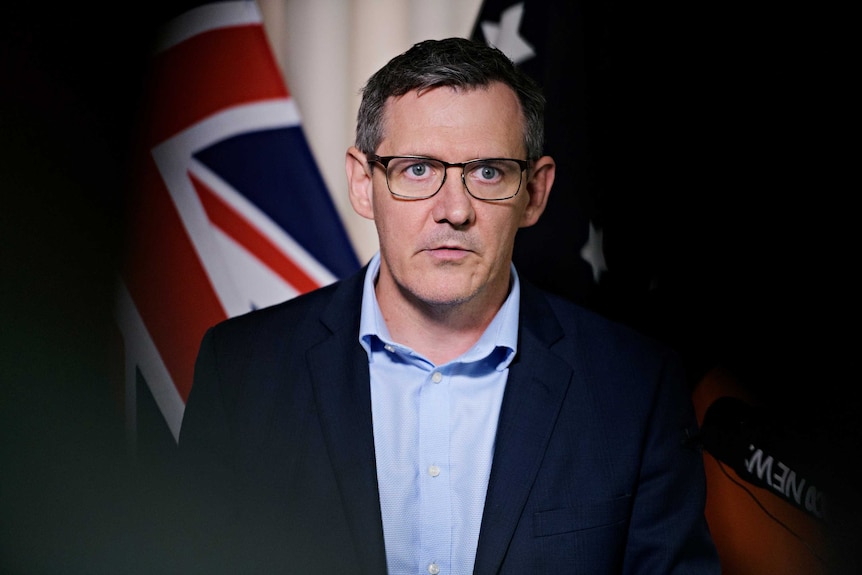 Chief Minister Michael Gunner is standing in front of a microphone with a serious expression. Behind him is the Australian flag.