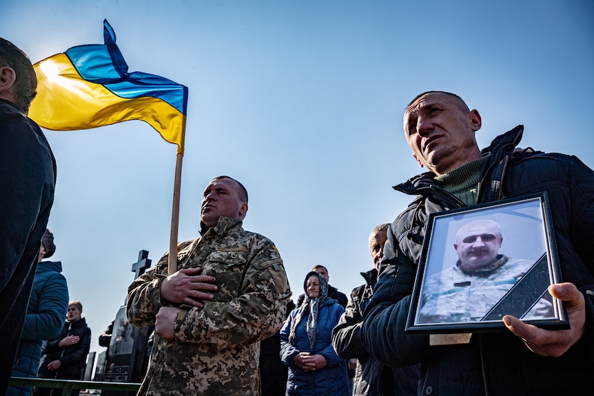 A framed photo of Bogdan is held by a mourner who is standing next to a soldier with the Ukrainian flag. 