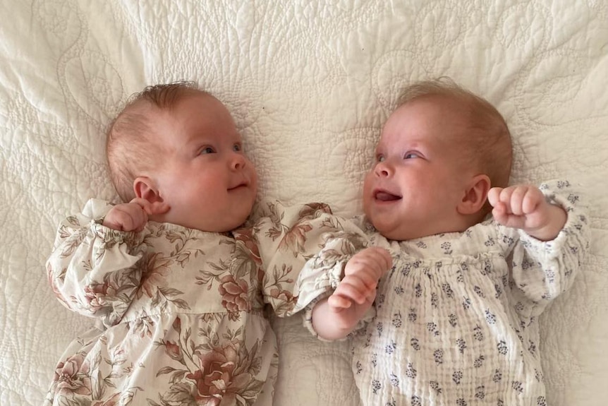 Two babies lying on a bed facing each other and smiling.