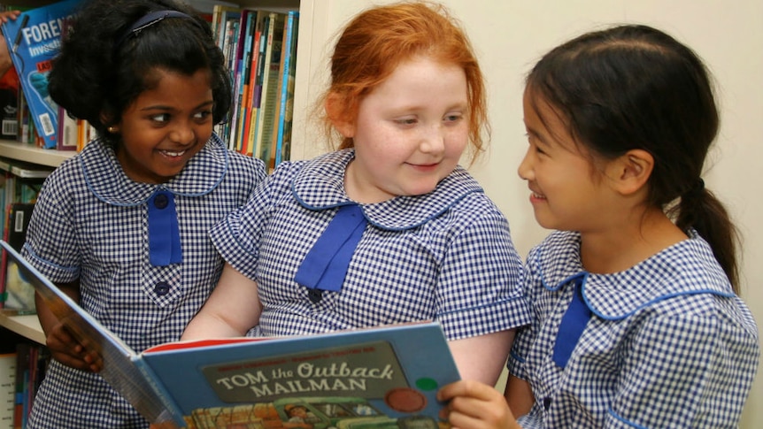Three young girls sitting next to each other in a library reading a book.