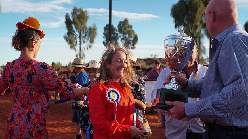 A smiling woman wearing an orange shirt and a winner's rosette is presented with a cup.