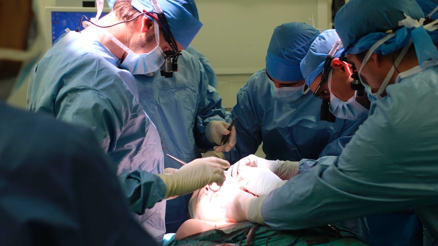 Surgeons in blue scrubs work on a a patient's head under a bright light