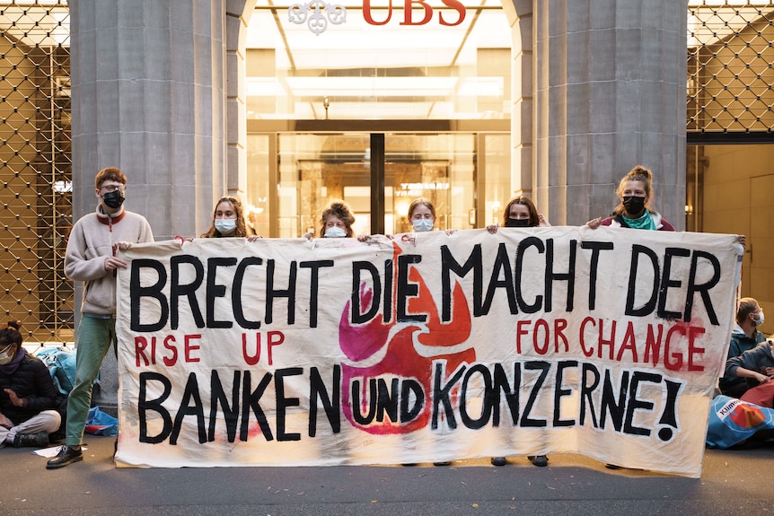 Six young people wearing face mask hold a white sign with German words on it in front of a bank building.