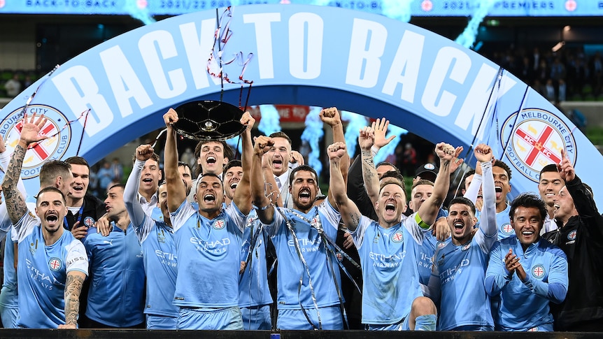 A soccer team wearing light blue celebrates with a 'back to back' sign and streamers while holding a trophy