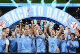 A soccer team wearing light blue celebrates with a 'back to back' sign and streamers while holding a trophy