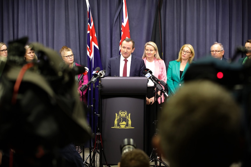 Long shot with cameras in the foreground of Mark McGowan at a podium