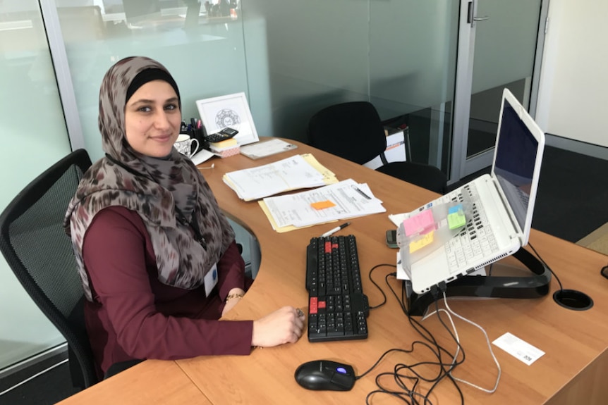 A smiling woman wearing a hijab sits at a tidy desk