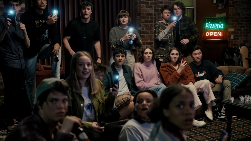 A group of teens in the movie Talk To Me hold up phones, filming something off-camera.