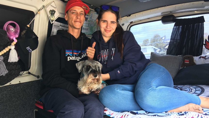 A man and a woman (and their dog) sit on a bed in a campervan.