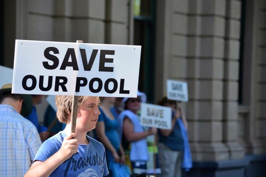 A teenage boy holding up a placard that says "SAVE OUR POOL".