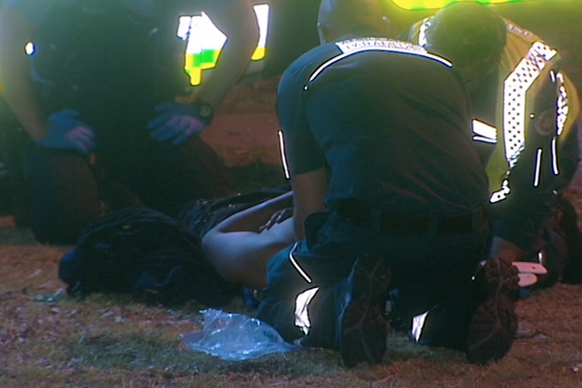 Three paramedics kneel on the ground outside treating a man lying on the ground.