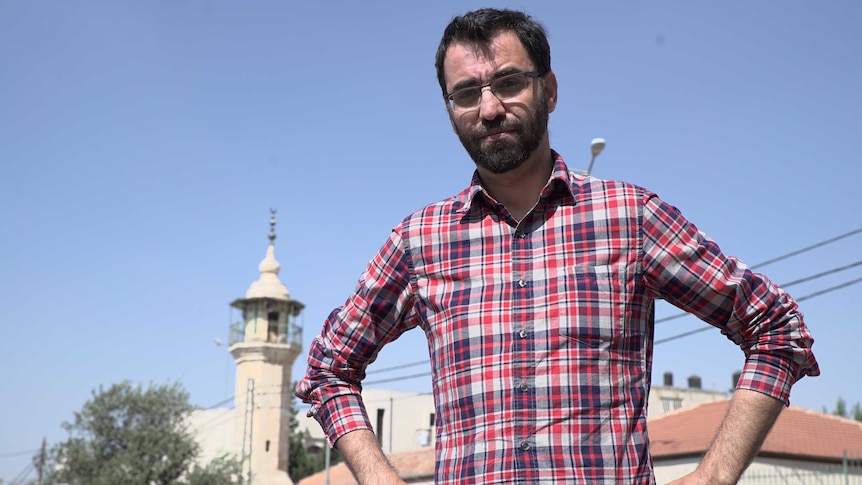 Aziz Abu Sarah stands with his hands on his hips, looking slightly down at the camera, standing outside a mosque at daytime
