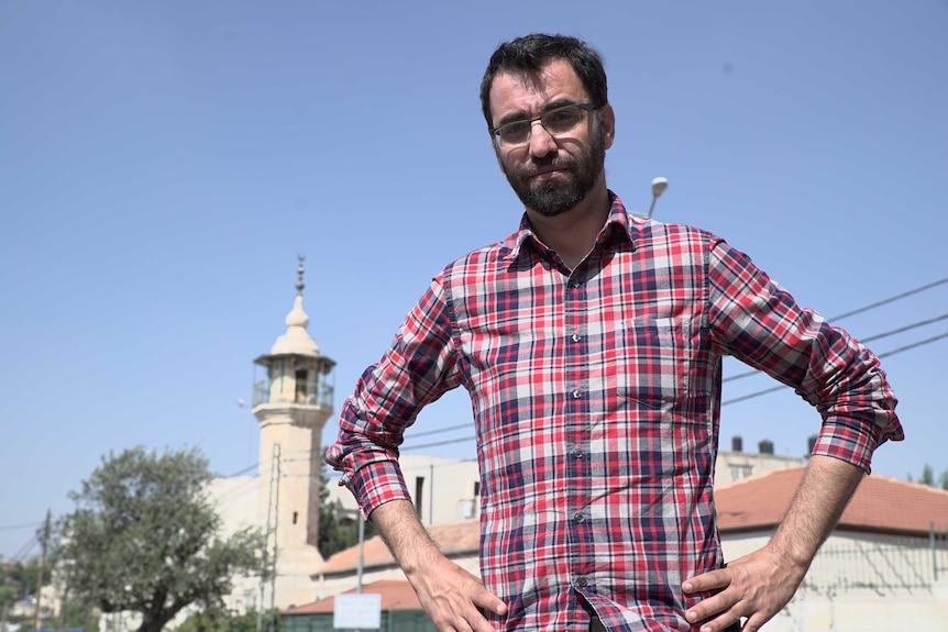 Aziz Abu Sarah stands with his hands on his hips, looking slightly down at the camera, standing outside a mosque at daytime