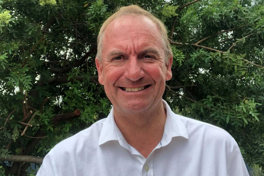 A man stands in a white shirt near a tree, smiling.