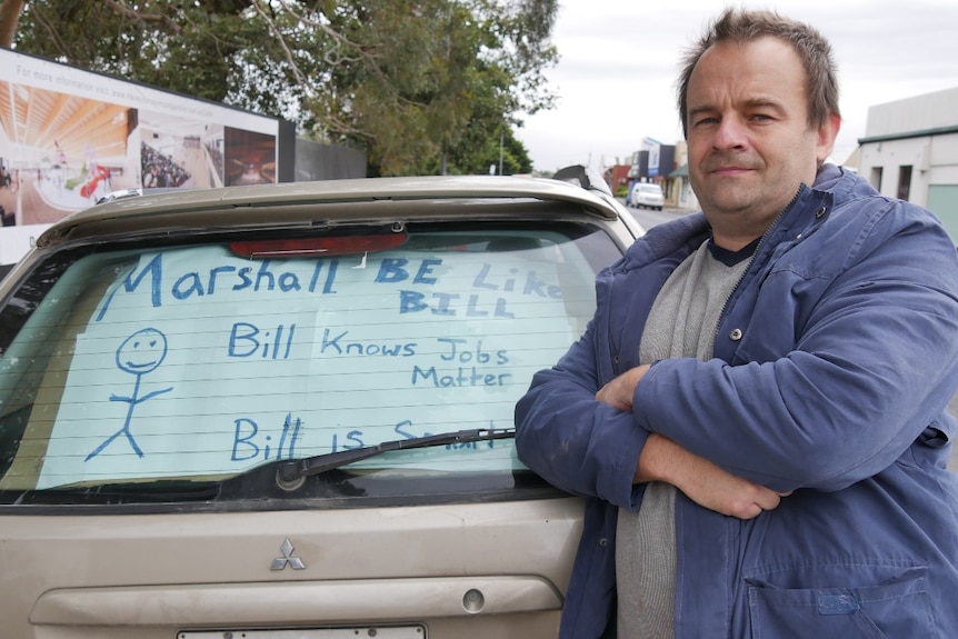 Man stands along car with protest sign in the window.
