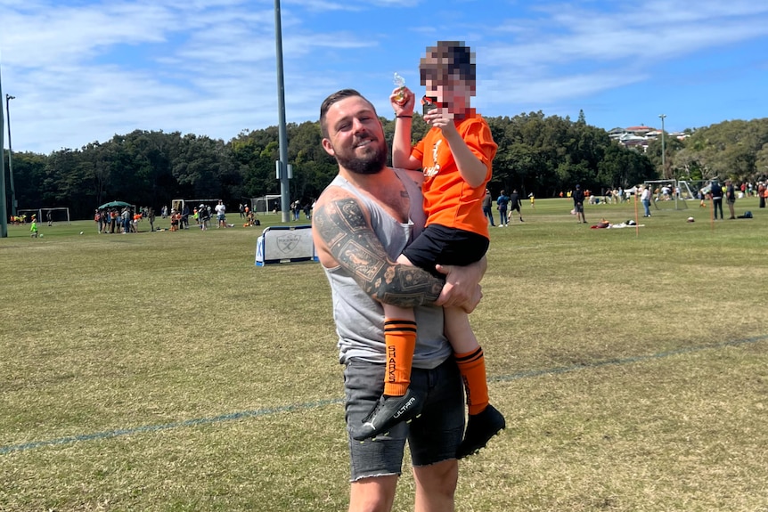 Young dad with tattoos holding son on soccer field on beautiful blue sky day.
