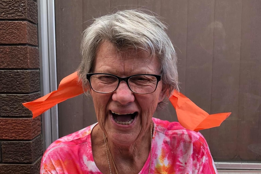 A laughing, elderly woman in a bright orange top with a stiff orange ribbon poking out from behind her head (on both sides).