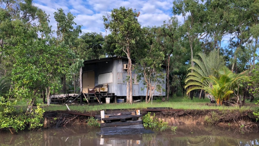 A hut sits on the banks of a river. Corrugated iron and silver surrounded by trees and steps entering the water.