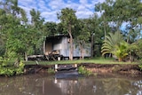 A hut sits on the banks of a river. Corrugated iron and silver surrounded by trees and steps entering the water.