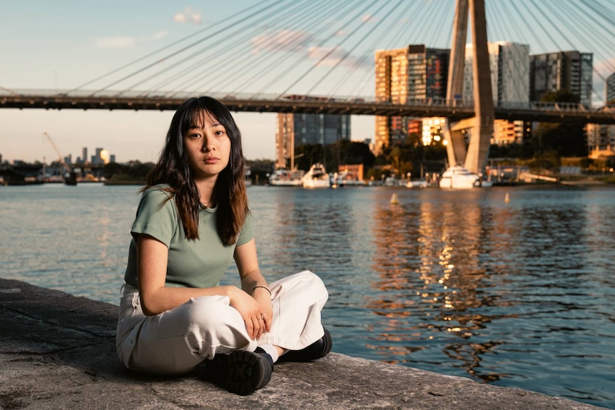 A woman sitting next to water with a bridge in the background