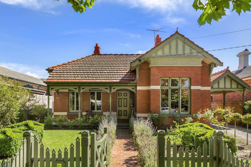 A historic double-fronted brick house sits behind a green picket fence and manicured front garden