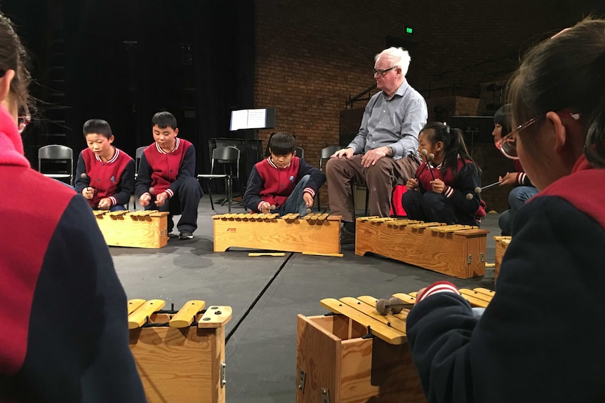 Children sit on the floor playing wooden xylophones while Richard Gill observes.