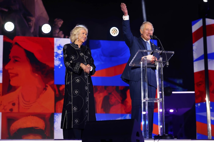 Camilla and Prince Charles on stage. Prince Charles lifts one hand up as he speaks into the microphone. 