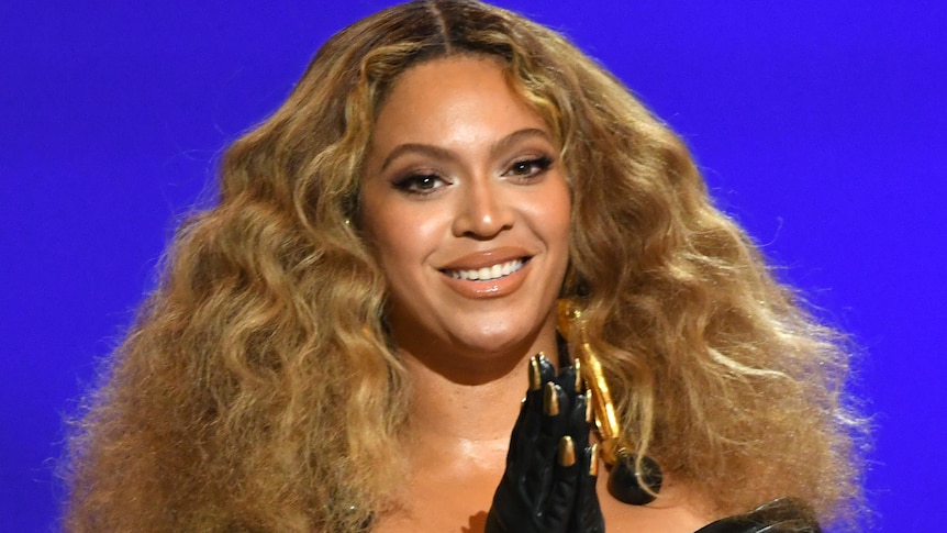 beyonce stands on stage clasping her hands together smiling wearing long black gloves