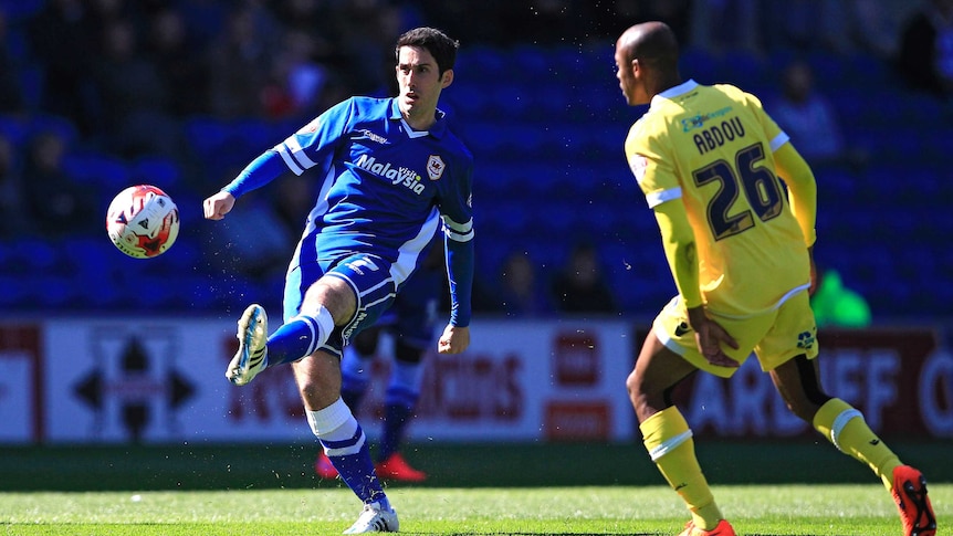 An English footballer looks towards goal as he hits the ball, while an opposing player watches.