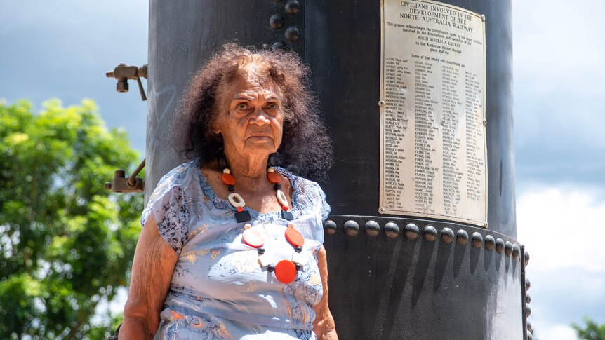 An elderly Indigenous woman stands in front of a memorial column on a bright day.
