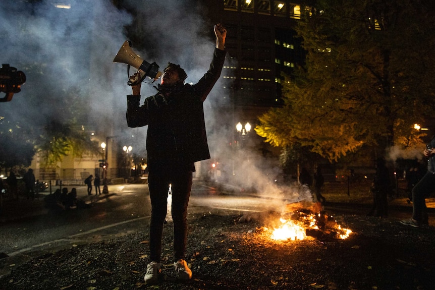 A black woman holding a megaphone raises her arm as a fire burns behind her in a smoky city park.