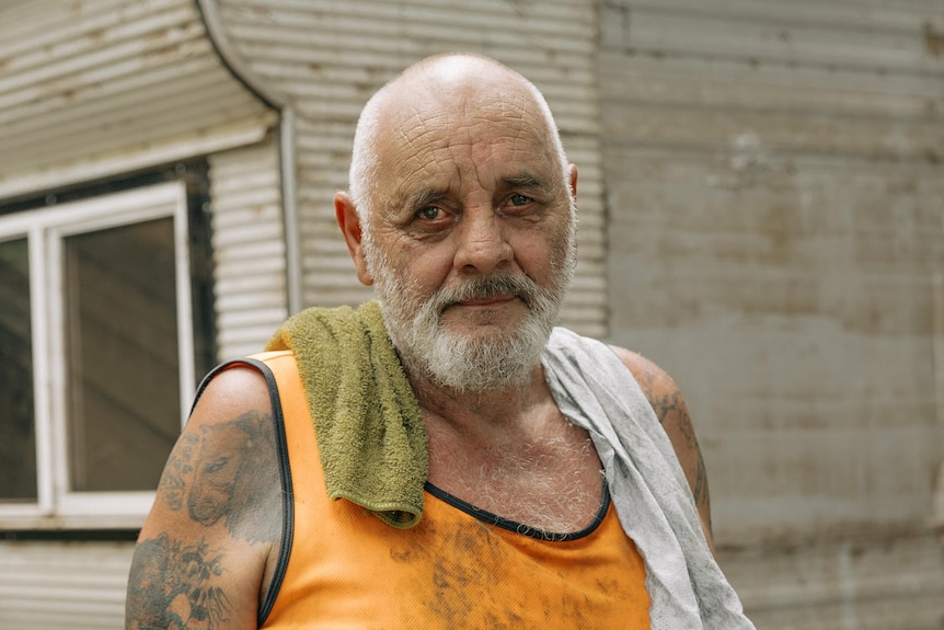 A muddy man in a singlet outside a dirty caravan