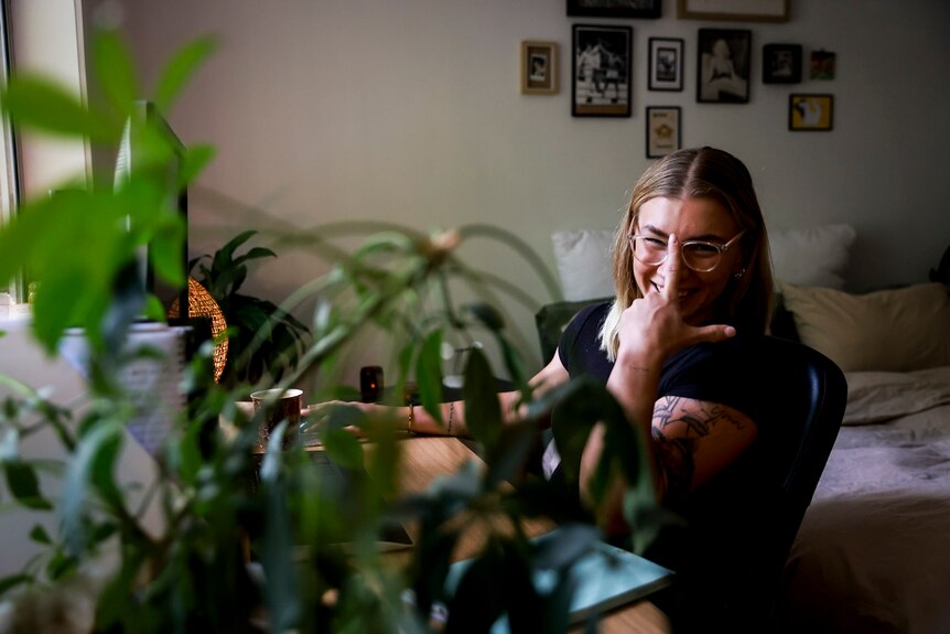 Marissa Williamson-Pohlman siting in their apartment, at a desk, with indoor plants and photos on the wall