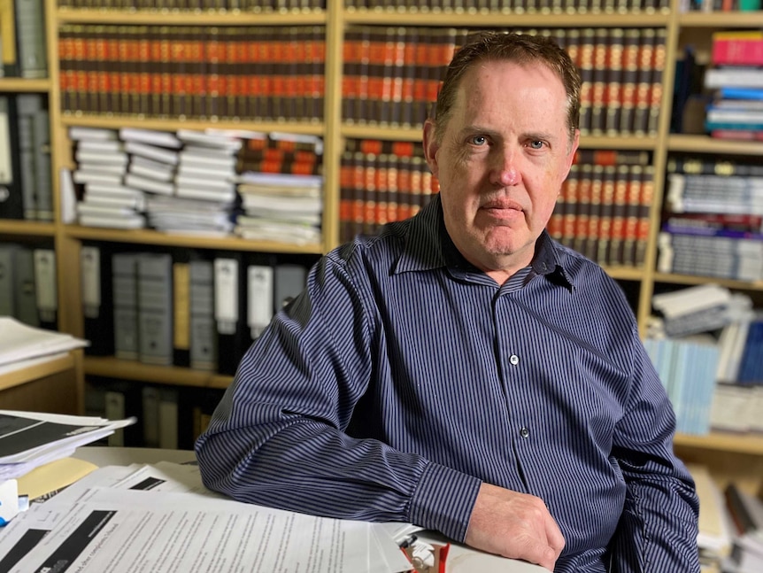 Professor Andrew Stewart is sitting at a desk full of papers in front of a shelf full of law books.