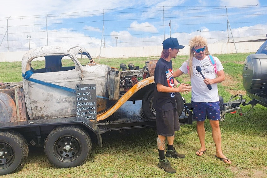 Two men stand in front of a burnt car on a trailer.