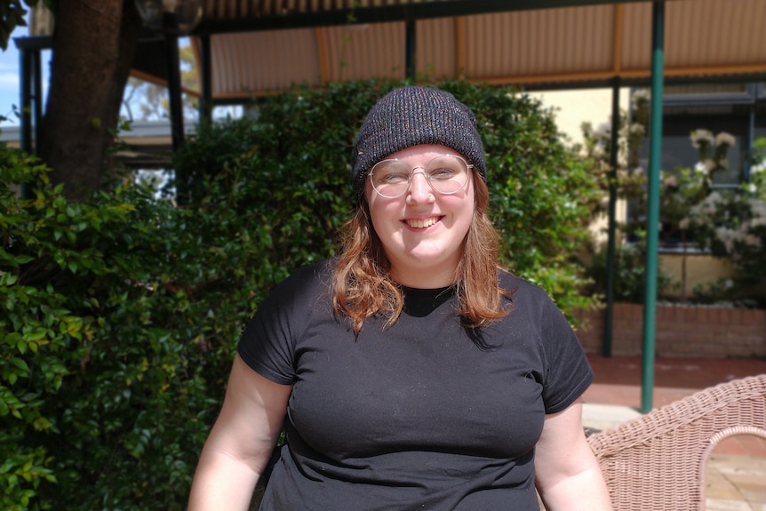A woman with glasses, a beanie, and a black shirt sitting down and smiling towards the camera. 