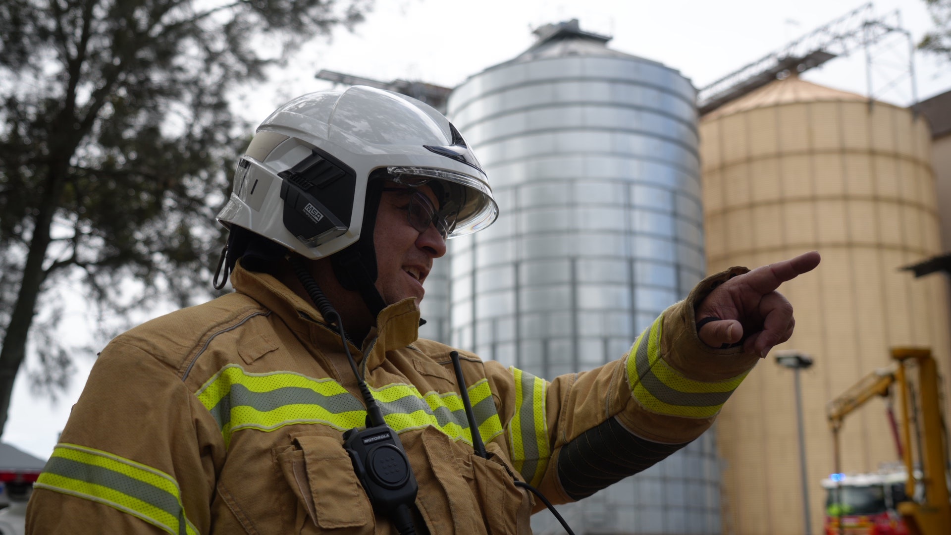 James Manuao in firefighting protective suit points with his hand. Silos are behind him.