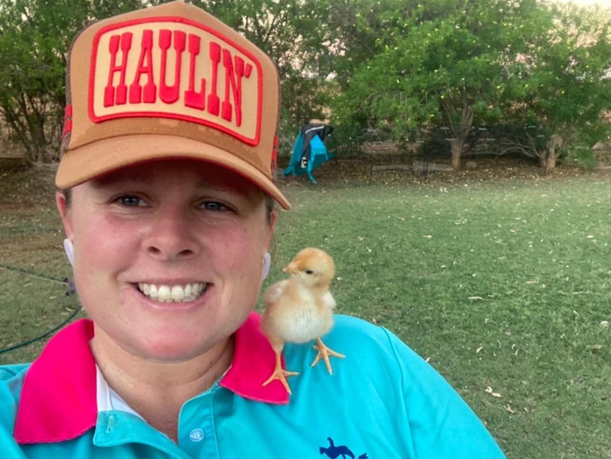 A smiling woman on a farm wears a hat that says "Haulin'". She has a chicken hatchling on her shoulder.