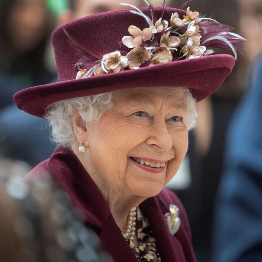 An elderly woman wearing a matching jacket and hat smiles as she talks with people.