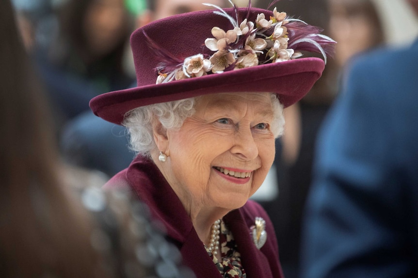 An elderly woman wearing a matching jacket and hat smiles as she talks with people.