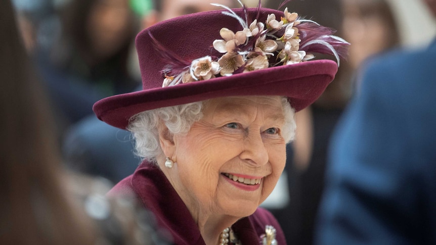 An elderly woman wearing a matching jacket and hat smiles as she talks with people.