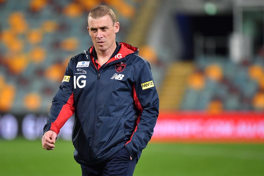 Melbourne AFL coach Simon Goodwin walks across a ground with one had in the pocket of his jacket.