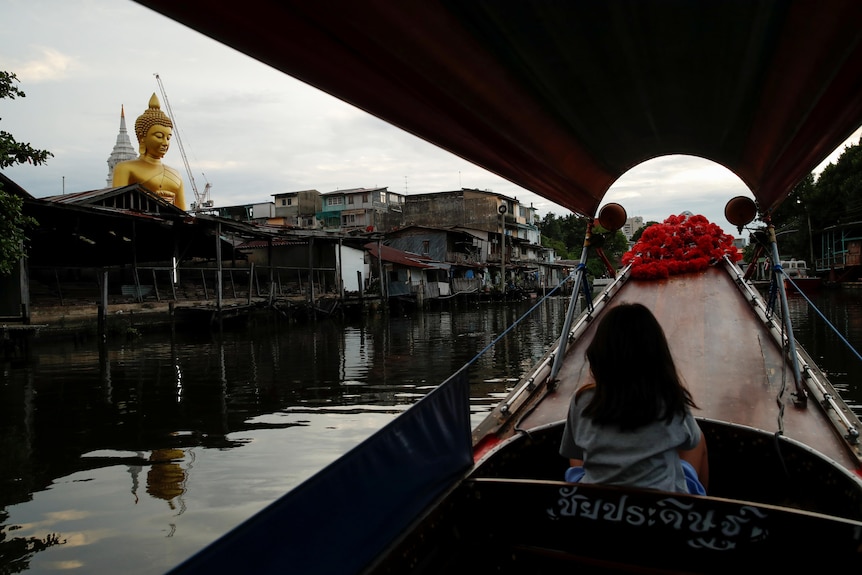 A girl sits in the front seat of a canal boat with roses on the bow, as it glides down a canal past a giant gold Buddha statue