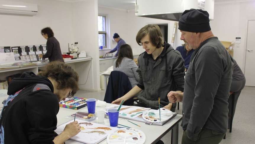 Julian Hume stands in a classroom advising two students on their paintings.