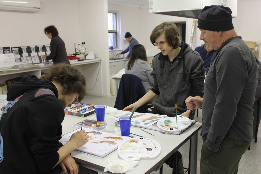 Julian Hume stands in a classroom advising two students on their paintings.