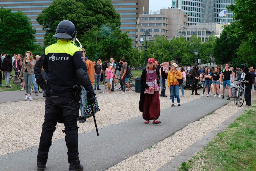 A policeman in a helmet stands facing a crowd, with a woman in the forefront.