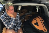 A young woman sits in the boot of her car which is packed with all of her belongings.