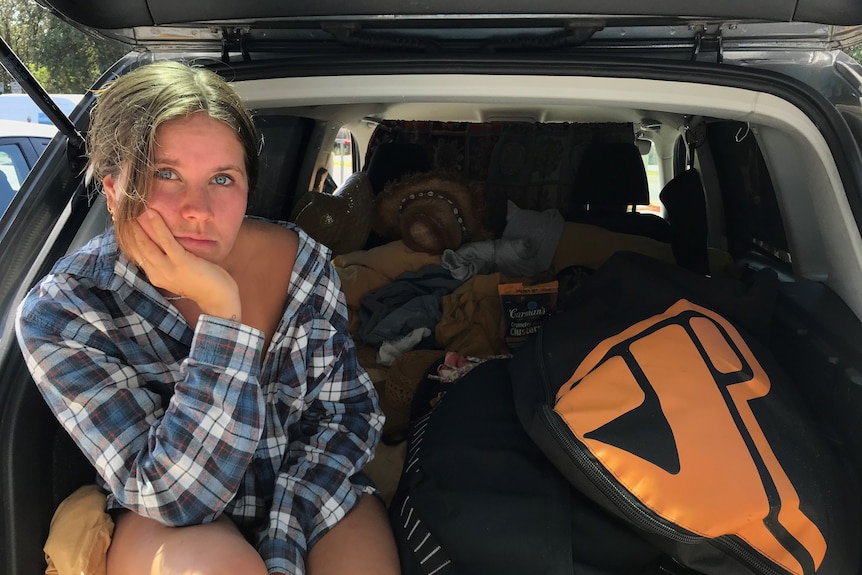 A young woman sits in the boot of her car which is packed with all of her belongings.